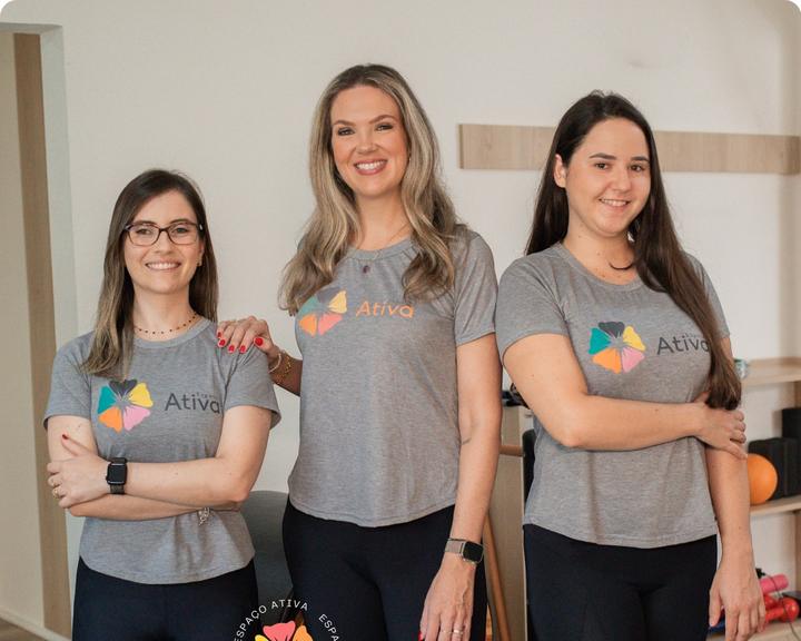 Alis, Júlia e Juliana lado a lado na sala de Pilates, de camiseta cinza da clínica