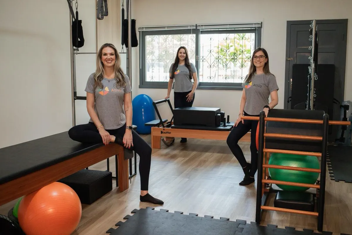 Júlia sentada no cadillac, Juliana em pé ao fundo junto ao reformer e Alis apoiada no espaldar, as três de camiseta cinza da clínica