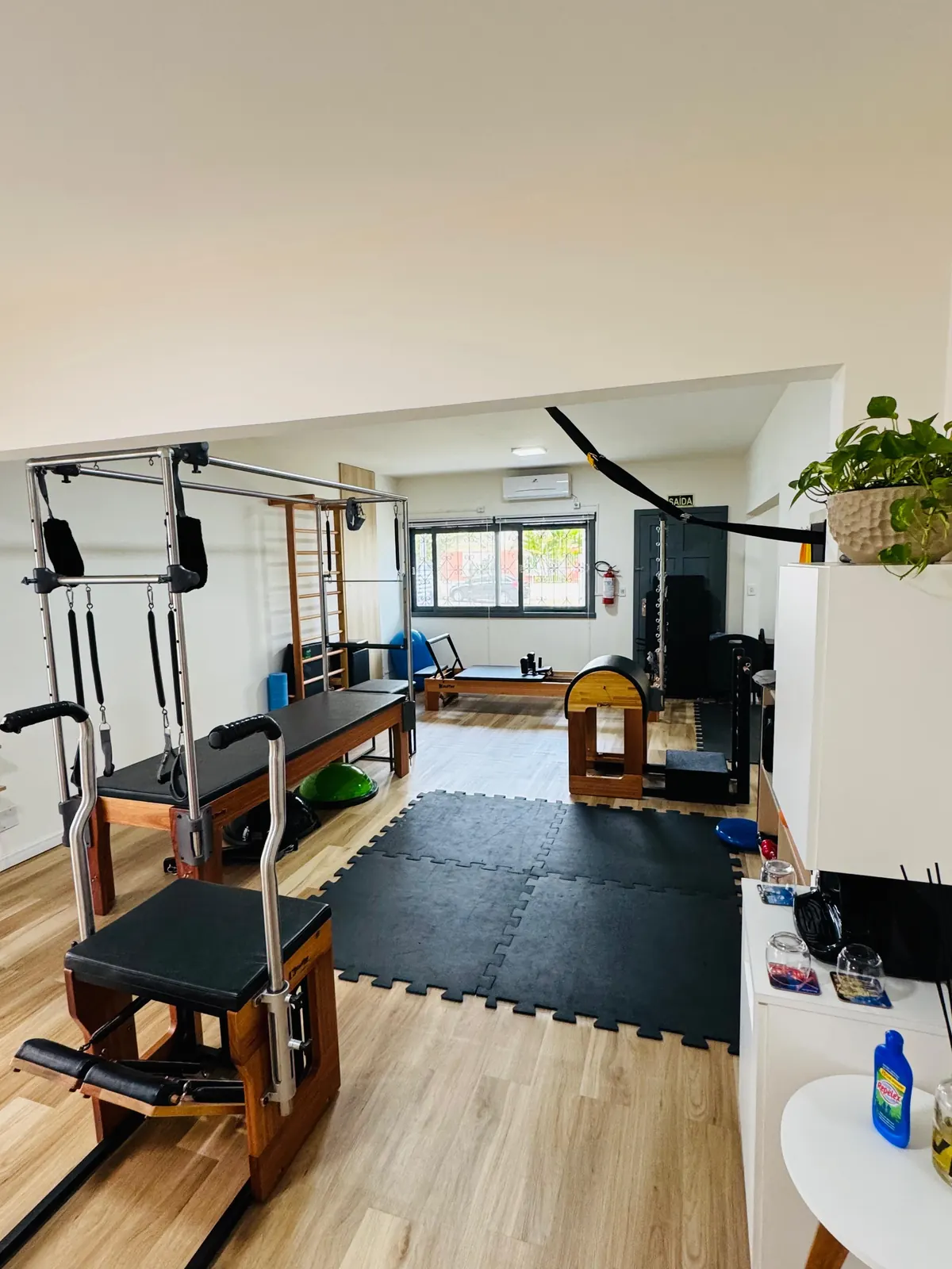 Sala de Pilates ampla: cadillac e chair em primeiro plano, espaldar e reformer ao fundo, tatame preto no centro e chão de madeira
